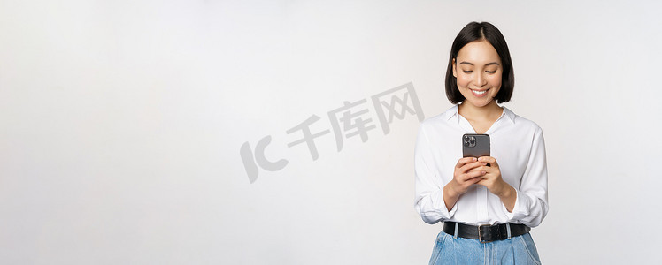 时尚的现代亚洲女孩使用手机应用程序，用手机聊天，微笑着，站在工作室背景下的白色衬衫