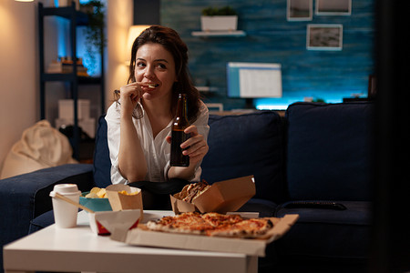 女人一边放松地坐在沙发上看电视，一边喝精酿啤酒和吃薯片