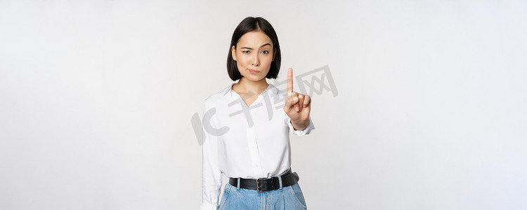 不。年轻、严肃、自信的亚洲女性表现出停下来、一根手指的手势、禁忌标志、站在白色背景上
