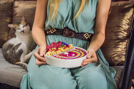 年轻女子坐在沙发上吃地中海早餐，吃健康的热带早餐、热带水果冰沙碗，装饰着彩色酸奶、姜黄和螺旋藻的图案。