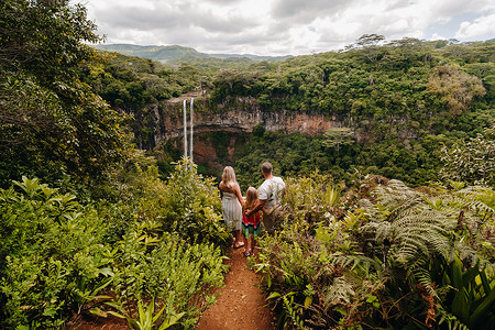 悬崖边上的公路摄影照片_毛里求斯岛查马雷尔公园瀑布附近的悬崖边上，有一个男人、一个女人和一个女儿。一对夫妇带着他们的女儿在毛里求斯岛的丛林里看着瀑布落下