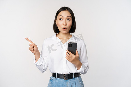 兴奋的亚洲女孩在移动应用程序上购物，拿着手机，用手指指着空的复制空间，显示促销文字，站在白色背景下的智能手机上