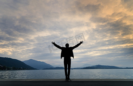 一个背对着镜头的男人的剪影，双臂在日落时分的湖景前伸展，天空中云彩被阳光染上色彩。