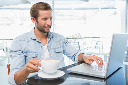 男人拿着咖啡摄影照片_年轻快乐的男人拿着咖啡在笔记本电脑上打字