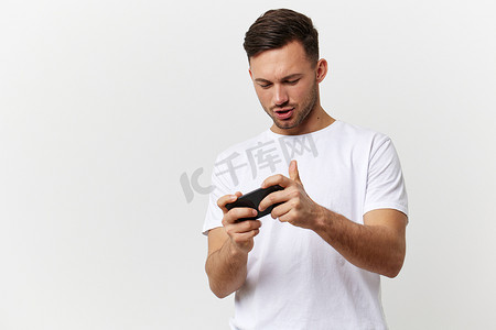 在线游戏摄影照片_穿着基本 T 恤、快乐、热情、晒黑的英俊男子玩在线游戏，手机在白色工作室背景下摆出孤立的姿势。