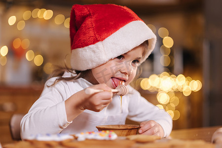 戴着红色圣诞老人帽子的小女孩用蜂蜜北斗七星吃蜂蜜，厨房装饰着童话般的灯光。