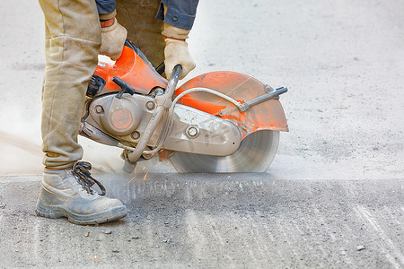 建筑工人用便携式汽油切割机和金刚石切割盘切割旧沥青，产生一团灰尘和火花。