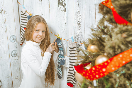 挂圣诞袜摄影照片_穿着白色毛衣、长着金色长发的可爱白人女孩挂着圣诞袜。