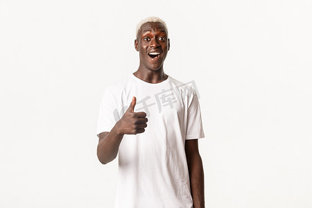 恭维摄影照片_令人印象深刻的微笑的非洲裔美国金发男子的肖像，竖起大拇指，称赞好的选择，恭维，白色背景