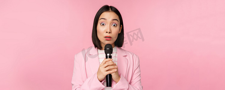 年轻的亚洲女售货员，穿西装的办公室女士，拿着麦克风，对着镜头感到震惊，说话，发表演讲，站在粉红色的背景上