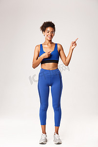 全长迷人的微笑非洲裔美国女性，梳着头发，穿着蓝色运动服，手指指向右上角，展示健身促销，白色背景