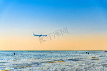 日落大海摄影照片_飞机在海面低空飞行，游客在水中游泳，日落时清澈的蓝橙色天空，飞机准备降落