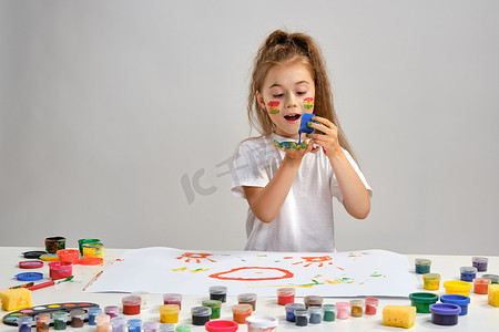 穿着白色 T 恤的小女孩坐在桌子旁，上面涂着什么，脸上和手上都涂着油漆。