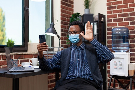手机病毒摄影照片_戴着科维德面具的初创公司员工挥手致意，在使用智能手机的在线采访中展示自己