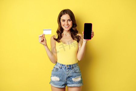 年轻美女模特展示智能手机空手机屏幕和信用卡，准备夏天，穿着背心和短裤，站在黄色背景上
