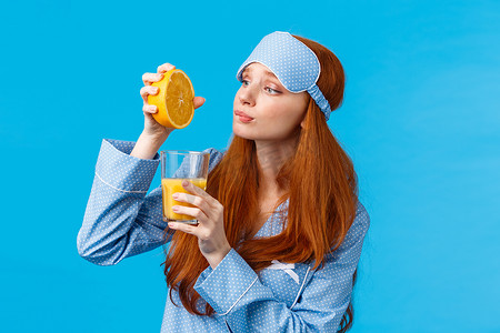 戴着睡眠面罩和睡衣的愚蠢可爱的红发白人女孩，看上去充满希望和渴望的表情，就像吃水果，在玻璃里挤橙子做早餐汁，蓝色背景