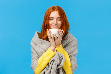 女孩享受着一杯热咖啡，闭着眼睛，高兴、高兴的微笑，裹着温暖的灰色围巾，在冬季低温天气中变暖，蓝色背景