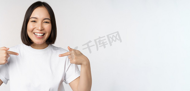 315徽标摄影照片_快乐而自信的亚洲女性，学生微笑着指着自己，自我推销，在 T 恤上展示徽标，站在白色背景上