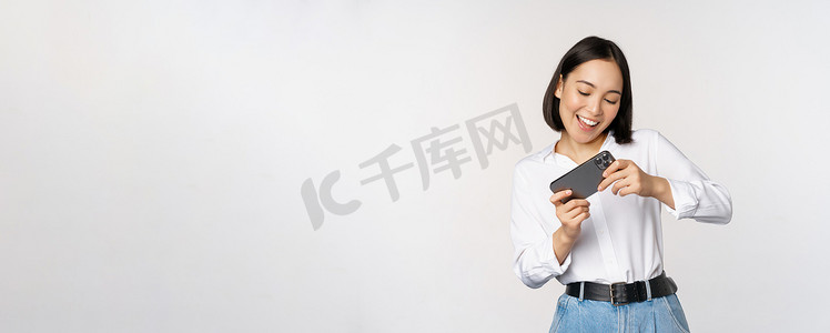 年轻的韩国女性，亚洲女孩在智能手机上玩移动视频游戏，看着水平手机屏幕，站在白色背景上