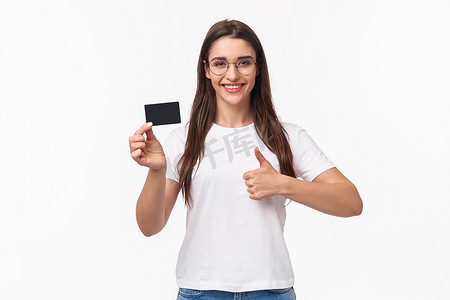 穿着 T 恤和眼镜的高兴、迷人的黑发年轻女性的肖像，推荐银行，出示信用卡，微笑满意地竖起大拇指，批准喜欢并建议网上购物