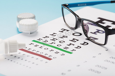 眼镜验光摄影照片_蓝色背景上带有隐形眼镜、滴剂和验光师眼睛测试图的眼镜。