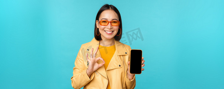手机界面摄影照片_微笑的韩国女性展示手机应用程序界面、智能手机应用程序、手机推荐、站在蓝色背景上