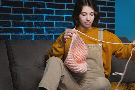 手工编织钩针摄影照片_有魅力的年轻女子坐在沙发上编织羊毛衣服。