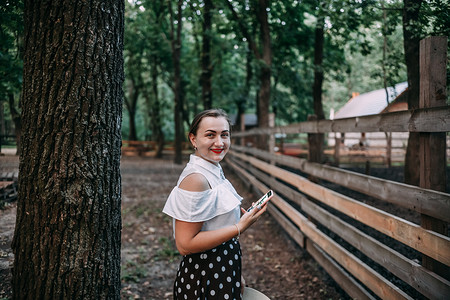 手机夏季夏季摄影照片_照片中，一位微笑的金发女郎在森林里拿着手机，他要拍照，这是一片美丽的夏季森林。