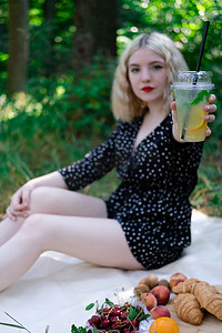漂亮的金发女孩在公园里穿着白色格子野餐，拿着一个塑料透明杯和柠檬水。