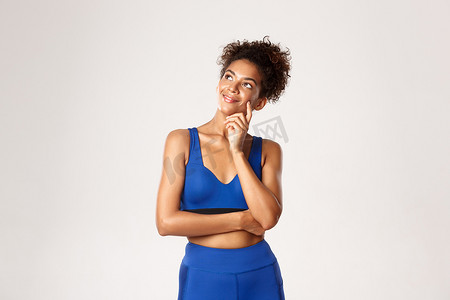 蓝色运动服摄影照片_工作室拍摄的非洲裔美国健身女性深思熟虑、面带微笑，身穿蓝色运动服，微笑着看着左上角，在白色背景下思考