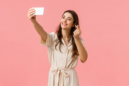 开朗可爱的年轻欧洲女性用滤镜拍可爱的自拍照，用手机摄像头拍照，微笑着在智能手机上展示韩国的心形手势，站在粉色背景上