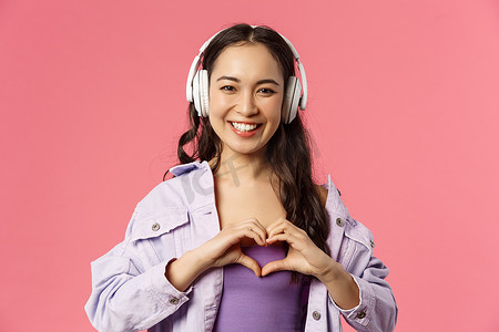 迷人、可爱的亚洲女孩在耳机里听音乐的特写肖像，在胸前展示心脏姿势，爱上她最喜欢的艺术家，歌手表演很棒的新歌，粉红色背景