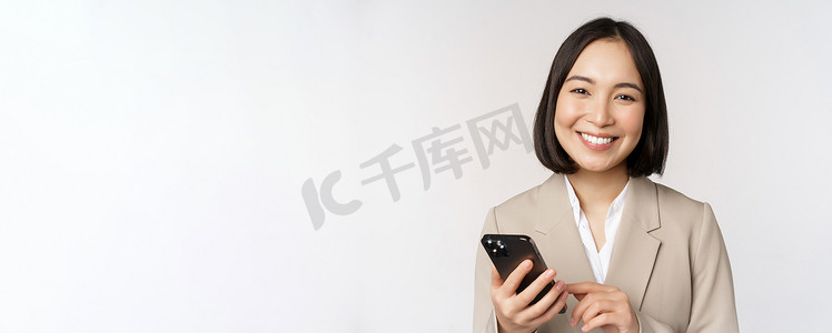 女士西装摄影照片_韩国女性、穿西装的企业女士的特写肖像，用手机微笑，拿着智能手机，站在白色背景上