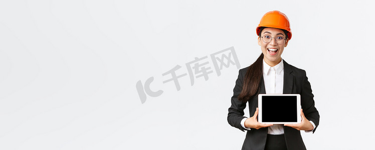 兴奋而惊讶的微笑亚洲女建筑工程师、身穿安全帽和西装的建筑师，展示数字平板电脑屏幕，表情惊讶，站在白色背景