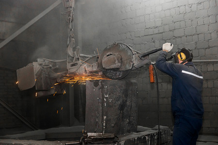 连衣裙子摄影照片_一名身穿工作连身衣和呼吸器的男子在工厂车间里用重型设备和磨石清洁铸铁管