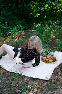 绿色格子布摄影照片_在公园里用格子布野餐的年轻女子的肖像，她正在读一本有美味小吃的书