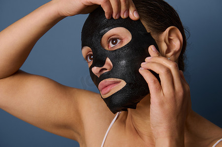 保湿补水摄影照片_美丽的女人在脸上戴上保湿、平滑、滋养、清洁的黑色织物面膜，隔离在带有复制空间的灰色背景中。