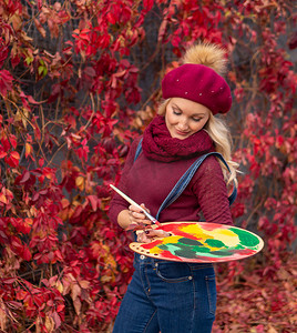 红色创意枫叶背景摄影照片_艺术家穿着蓝色牛仔裤和酒红色毛衣，用红色颜料在树叶上画上红色的枫叶。
