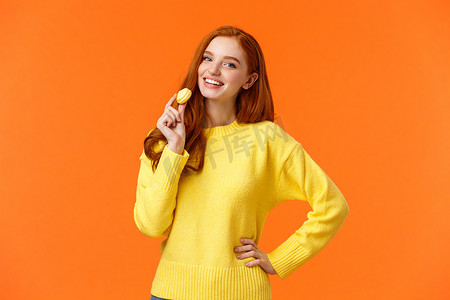 腰部肖像可爱的红发女孩，嘴边拿着美味的黄色马卡龙，高兴地微笑，喜欢吃糖果，为浪漫的情人节约会买甜点，摆出欢快的橙色背景