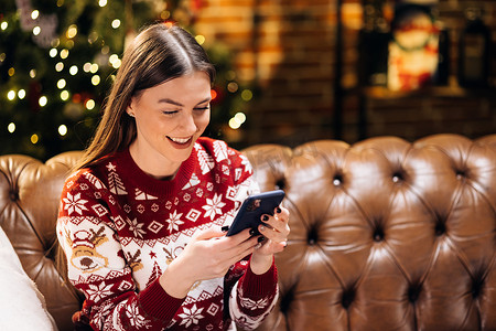 性格开朗的漂亮女人使用智能手机，玩在线游戏，在社交网络中交流，在线为朋友或家人发送圣诞祝福，在家圣诞树附近的沙发上休息
