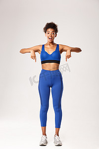 全长惊讶的非洲裔美国健身女孩，穿着蓝色运动服，手指向下，微笑着，站在白色背景下