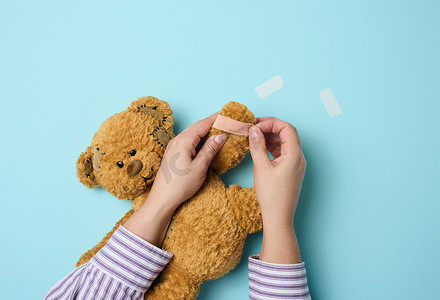 女手握着一只棕色泰迪熊，在蓝色背景上粘上医用胶布，电车治疗
