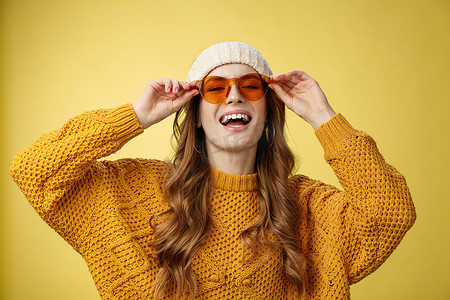 快乐时尚的白人女友享受阳光冬季滑雪胜地，戴着墨镜帽子温暖的毛衣，开心地笑着触摸眼镜，表达俏皮的快乐心情黄色背景