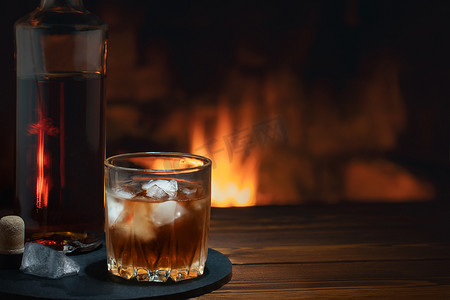 冰火摄影照片_燃烧的壁炉附近的桌子上放着一瓶和一杯加冰的威士忌。