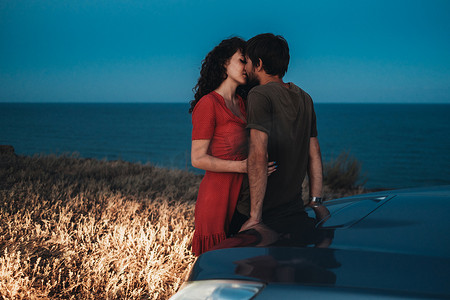黄昏时分，年轻夫妇在海岸线上的汽车附近亲吻和拥抱
