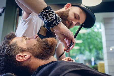 剃胡子的男人摄影照片_留着胡子的年轻人准备在熟练理发师的发廊里剃须