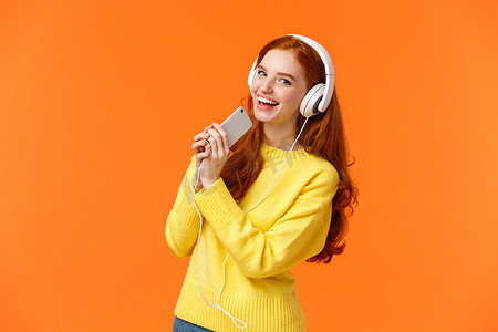 冬天的音乐背景摄影照片_漂亮的红发白人女性喜欢听音乐，戴着耳机在智能手机上玩卡拉 OK 应用程序，高兴地看着相机，拿着手机像麦克风，橙色背景