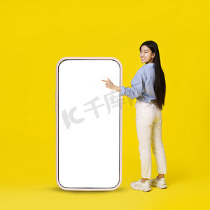 亚洲女孩指着巨大的智能手机，白色的空白屏幕站在后面，高兴地指着手指，穿着黄色背景的休闲装。