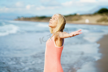 美丽的年长女性张开双臂在美丽的海滩上，度过她的闲暇时光。