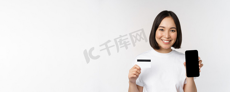 手机app购物摄影照片_站在白色背景上微笑的年轻亚洲女性的肖像，展示手机屏幕和信用卡